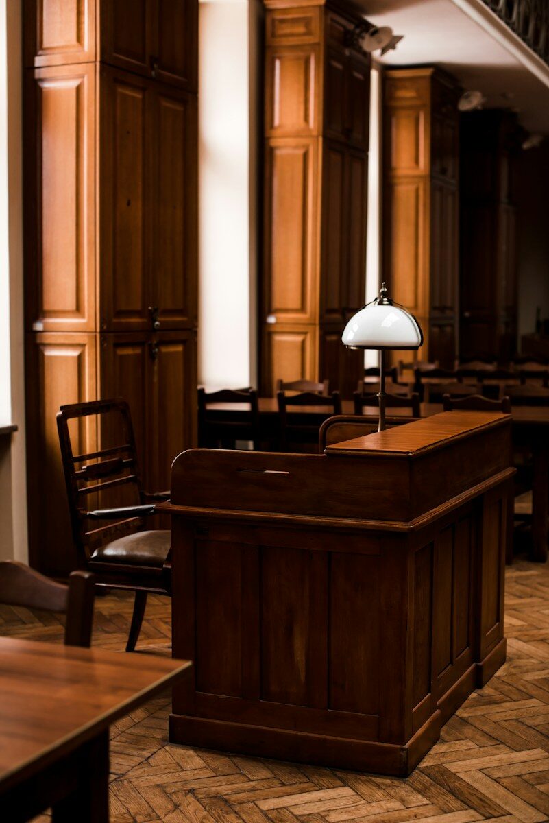 A wooden reception desk with a white lamp sits in a warm-toned, elegant library, surrounded by rows of chairs and tall cabinets.