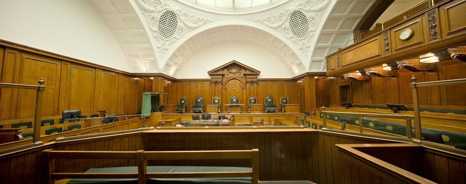 Wood-paneled courtroom featuring ornate architecture, with rows of green chairs for judges and a large decorative feature behind the judge's bench.