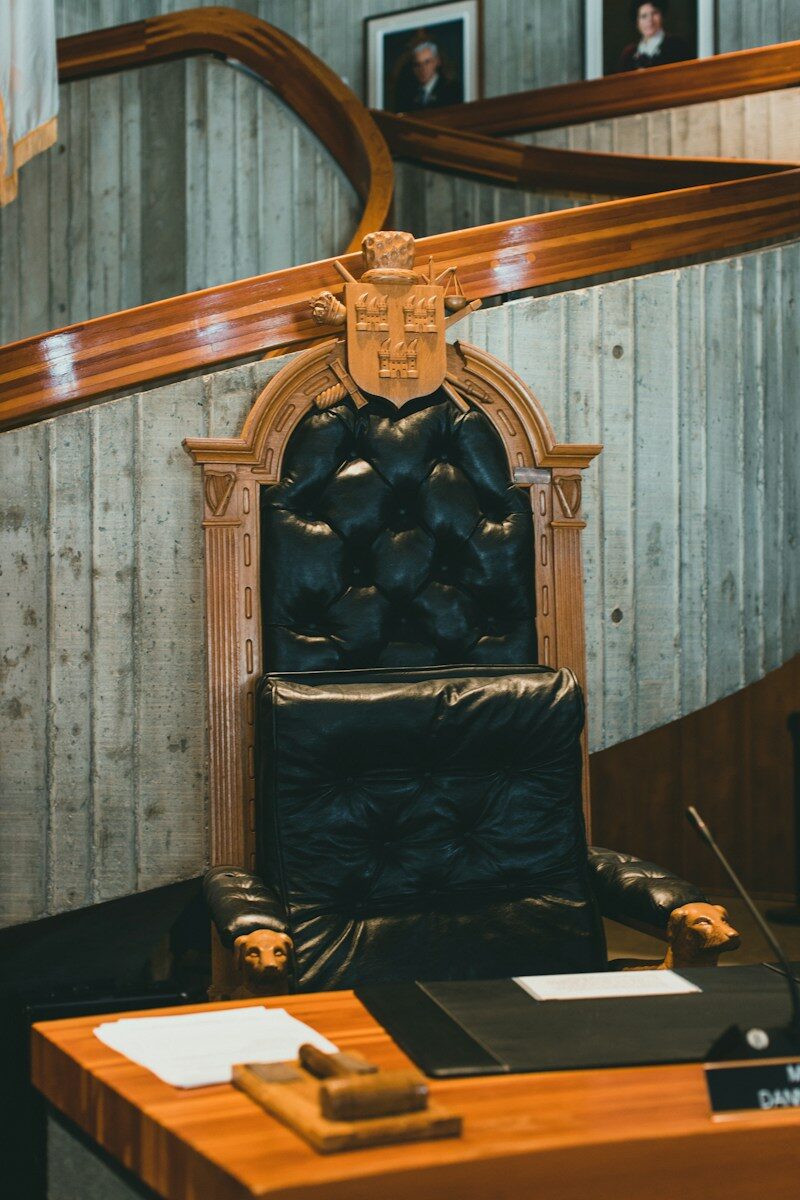 Black leather seat with wooden accents, placed at a desk with papers and a gavel, within a council chamber setting. Represents formal governance.