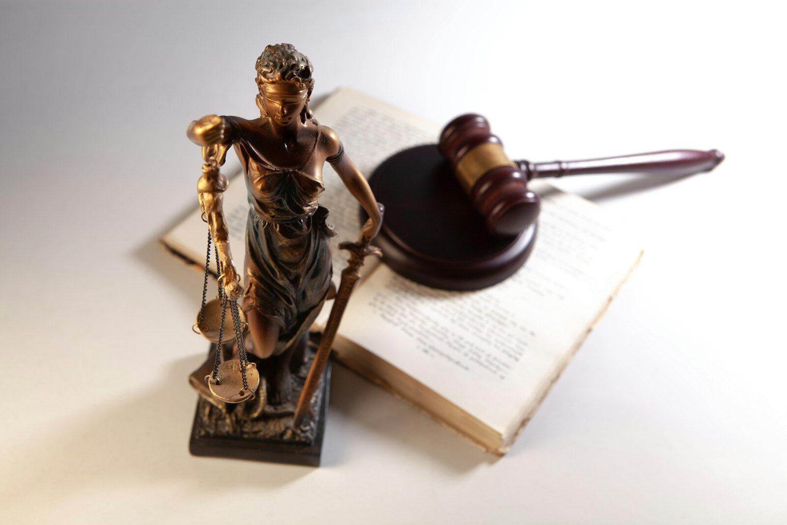 Bronze statue of Lady Justice, blindfolded and holding scales, next to a wooden gavel on an open law book. Represents legal equity and justice.